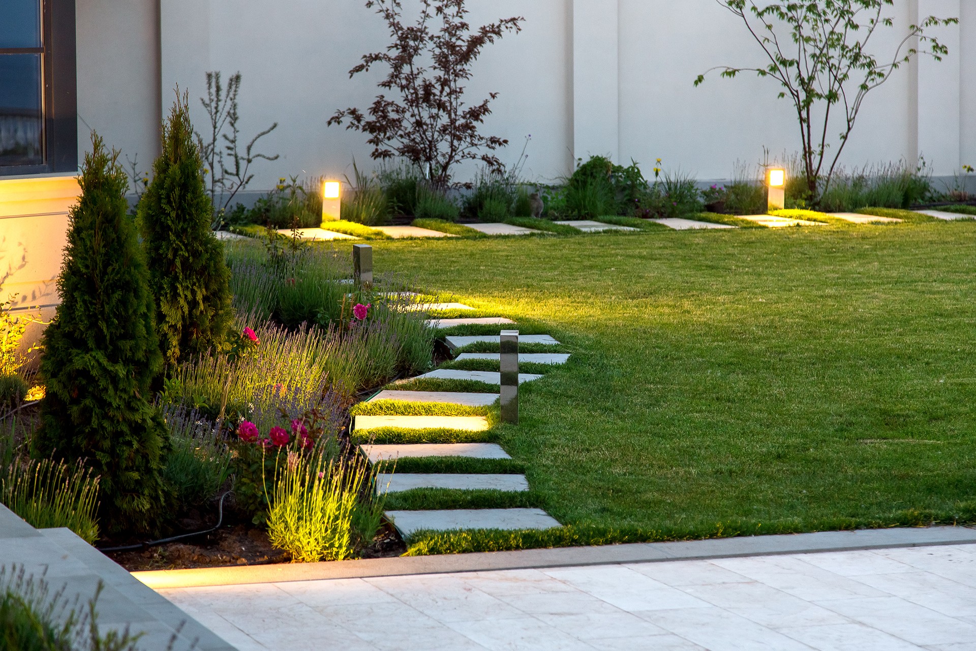 backyard of the mansion with a flowerbed and a lawn of green grass with a marble walkway of square tiles in the evening with a garden lighting with decorative ground lamps illuminating a warm light.